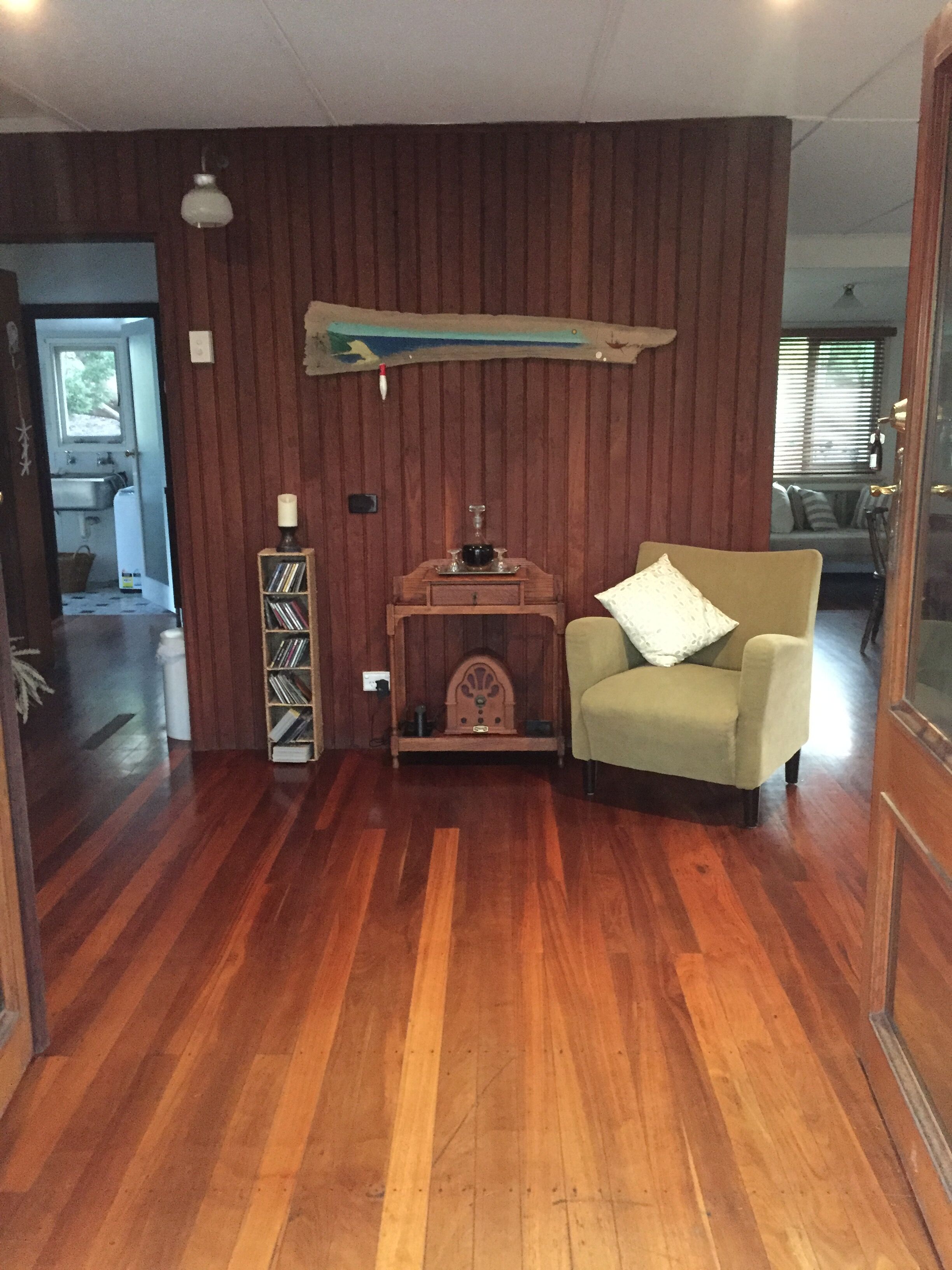 Une pièce aux murs lambrés, avec un plancher en bois, un fauteuil beige, une petite table d’appoint en bois sur laquelle se trouve une bougie, et une étagère murale remplie de livres.