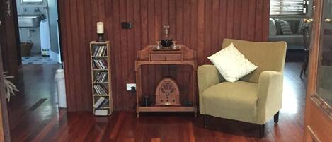 Une pièce aux murs lambrés, avec un plancher en bois, un fauteuil beige, une petite table d’appoint en bois sur laquelle se trouve une bougie, et une étagère murale remplie de livres.