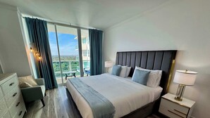 A hotel room with a large bed, a nightstand, a dresser, and a view of the city. - Balcony Ocean Views | Pool, Gym, Wi-Fi & Parking (Miami)