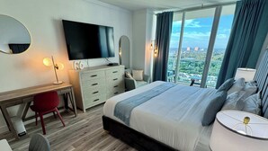 A hotel room with a large bed, a desk with a chair, a TV, and a view of the city. - Balcony Ocean Views | Pool, Gym, Wi-Fi & Parking (Miami)