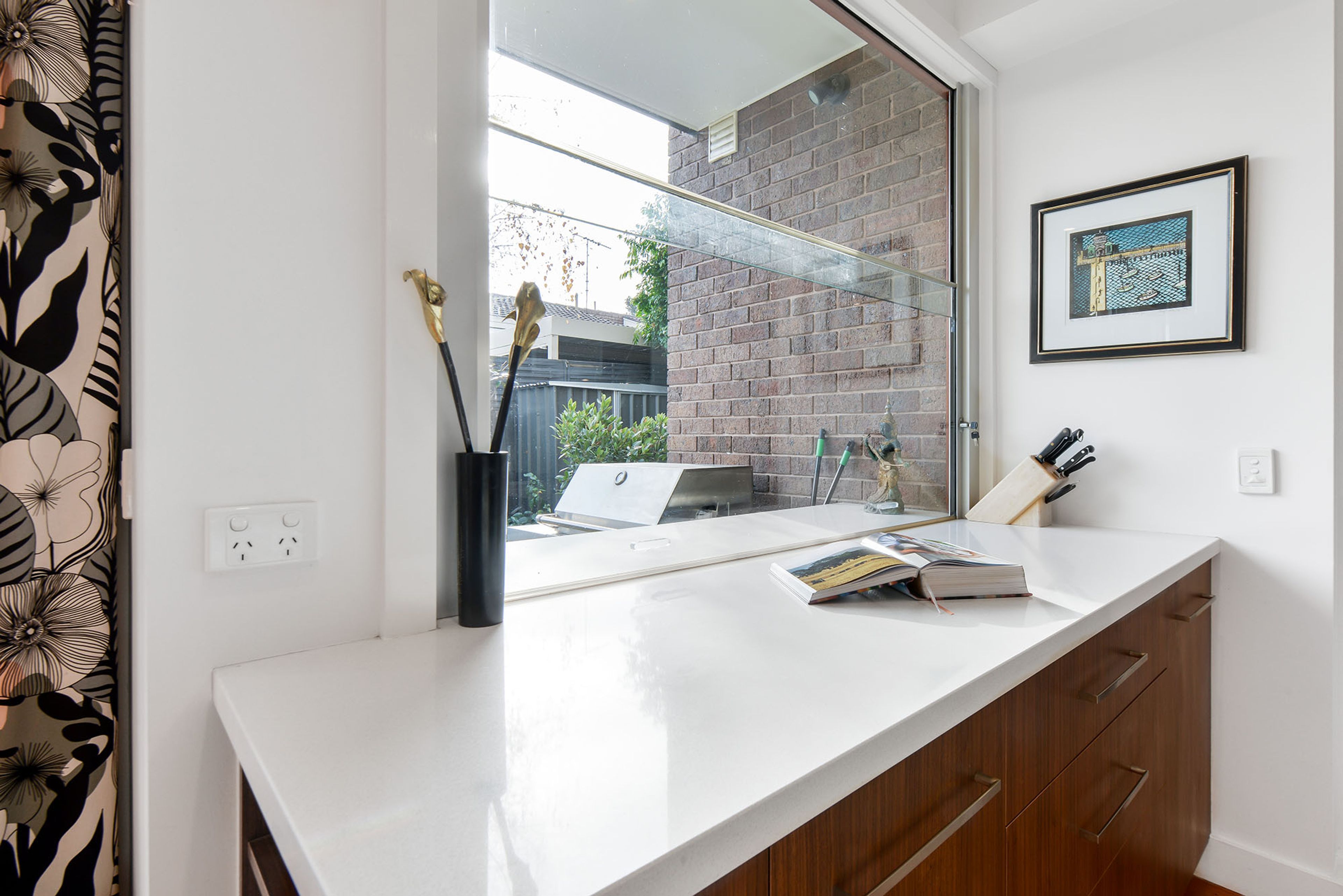 A modern kitchen with a white countertop, a window with a view of a brick wall, and a framed picture on the wall.