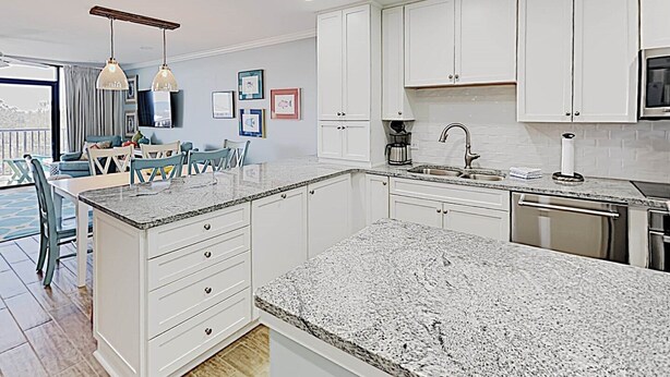 A modern kitchen with white cabinets, granite countertops, and a dining area with blue chairs. - Retreat 514 - 2nd Row, Great View, Pool, Ocean! (Pawleys Island)