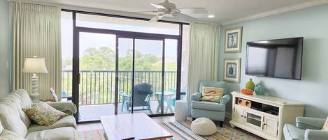 A living room with a sofa, armchairs, a coffee table, and a TV. There's a balcony with a view of greenery.