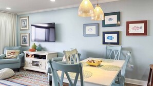 A living room with a TV, dining table, chairs, and artwork on the walls. - Retreat 514 - 2nd Row, Great View, Pool, Ocean! (Pawleys Island)