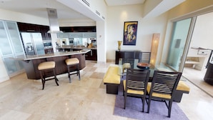 A modern kitchen with dark cabinetry, a central island with stools, and a dining area with a glass table and chairs. - Luxury Beachfront Condo! 3BD Condo for rent in Los Muertos Beach, Puerto vallart (Puerto Vallarta)