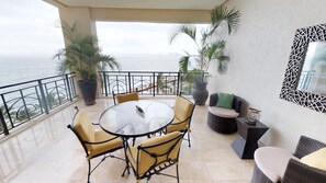 A balcony with a round table and chairs, potted plants, and a view of the sea.