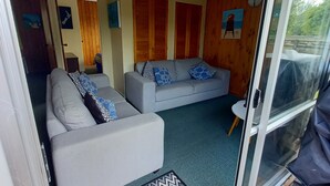 A living room with a grey sofa and a patterned rug, wooden panel walls, and a sliding glass door leading to an outdoor area. - Tui's Rest Whangamata- DOGS WELCOME, suitable for families (Whangamata)