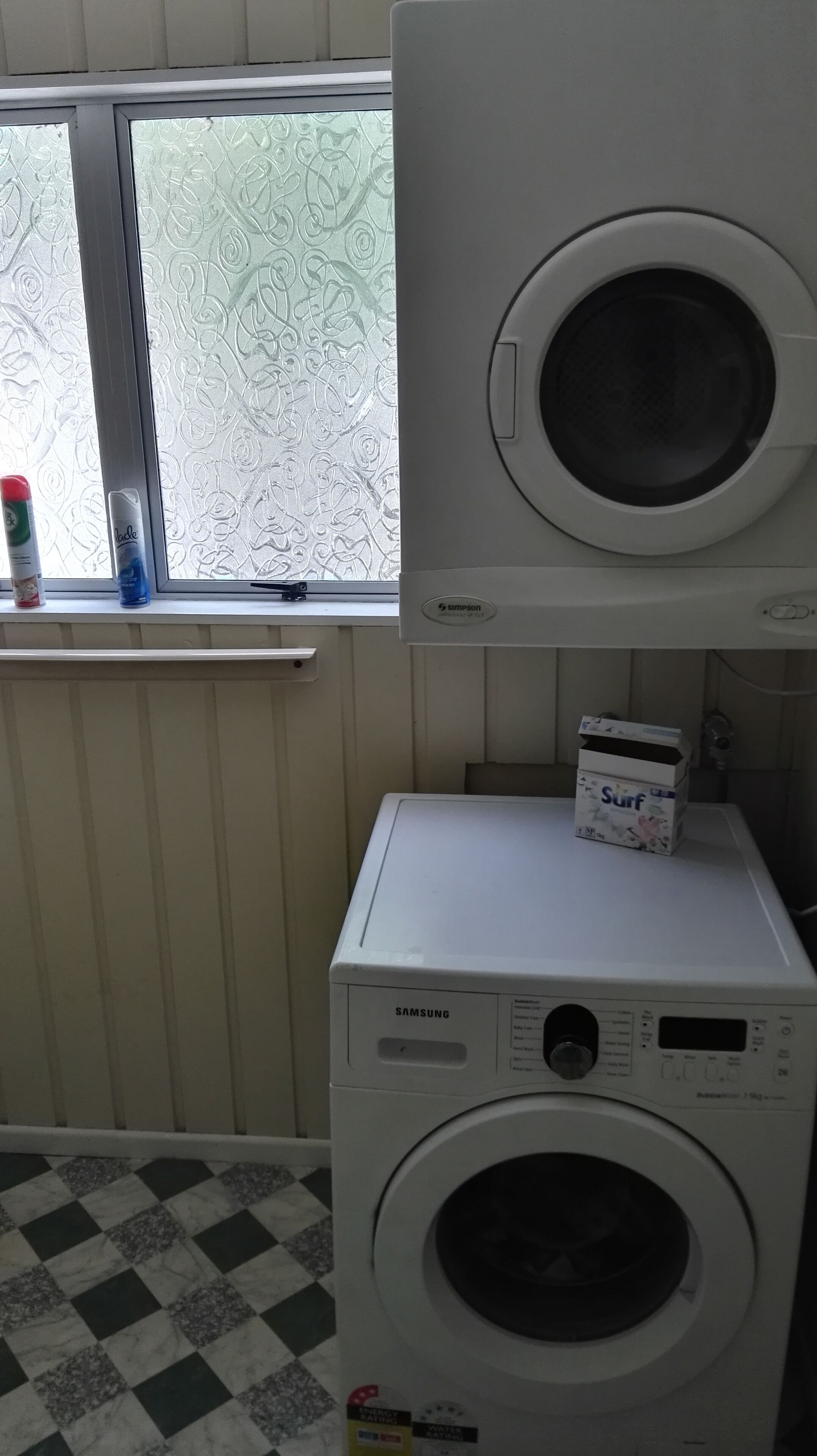 A laundry area with a Samsung washer and dryer stacked on top of each other, a box of Surf detergent, and a window with frosted glass.