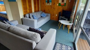 A living room with a grey sofa, a white armchair, and a small round table. There are wooden panel walls and a large window with a view of the outdoors. - Tui's Rest Whangamata- DOGS WELCOME, suitable for families (Whangamata)