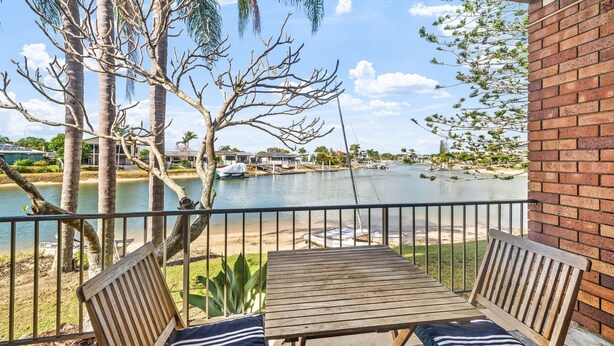 A wooden deck with a table and chairs, overlooking a body of water with boats and houses in the background. - Anjuna, 2 bedroom apartment on canal (Mooloolaba)