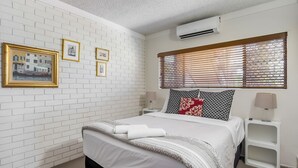 A bedroom with a white brick wall, a bed with white bedding, a window with blinds, and three framed pictures on the wall. - Anjuna, 2 bedroom apartment on canal (Mooloolaba)