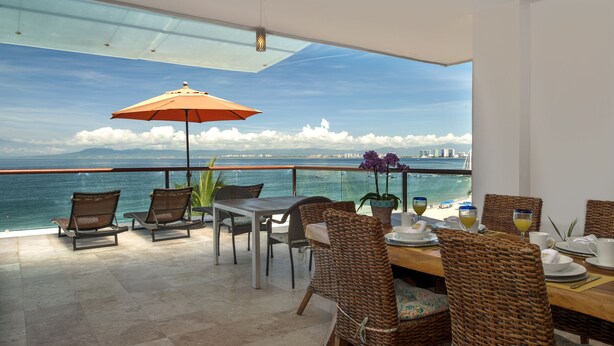 Uma área de refeições com vista para o mar e uma ponte, com cadeiras de vime, uma mesa com pratos e um guarda-sol.