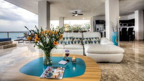Uma sala de estar moderna com um sofá branco, uma mesa de centro com revistas e velas, e vista para o mar.