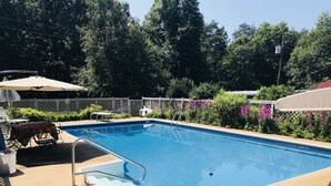 A rectangular swimming pool with a metal ladder, surrounded by a concrete deck. There are lounge chairs and a table with a tablecloth, and a fence with flowering plants.