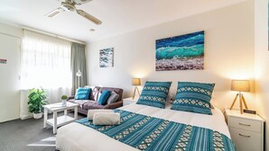 Ett hotellrum med en säng, en soffa, ett skrivbord, ett nattduksbord, en lampa, en klocka och en tavla som föreställer en strandvy.