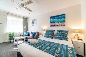 A hotel room with a bed, a sofa, a desk, a nightstand, a lamp, a clock, and a painting of a beach scene.