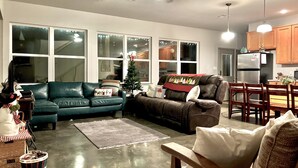 Una sala de estar con un sofá verde, un sillón reclinable marrón, un pequeño árbol de Navidad y un comedor con sillas de madera y una mesa.