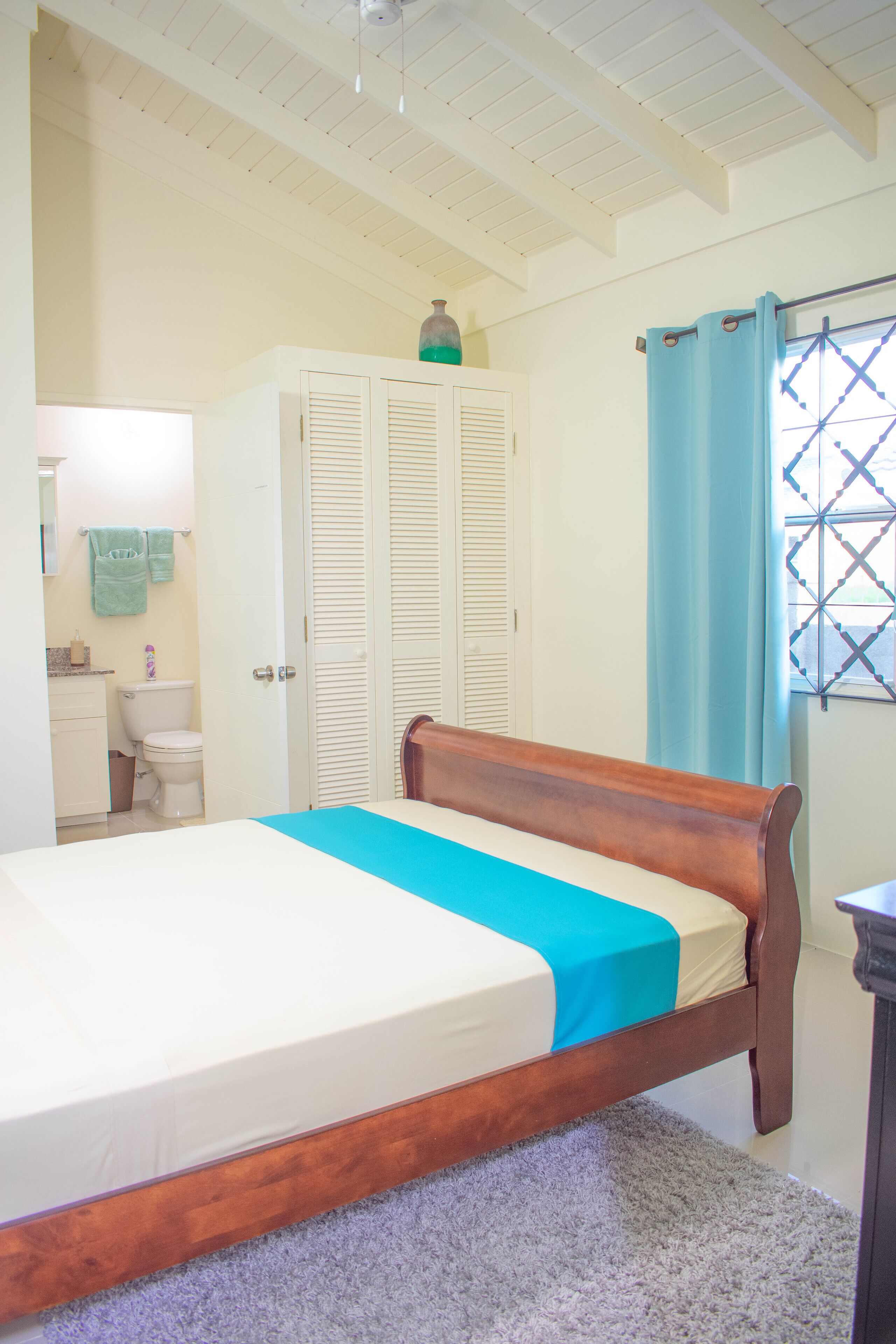 A bedroom with a wooden bed, a white wardrobe, a bathroom with a toilet, and a window with a curtain.