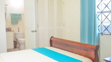 A bedroom with a wooden bed, a white wardrobe, a bathroom with a toilet, and a window with a curtain.
