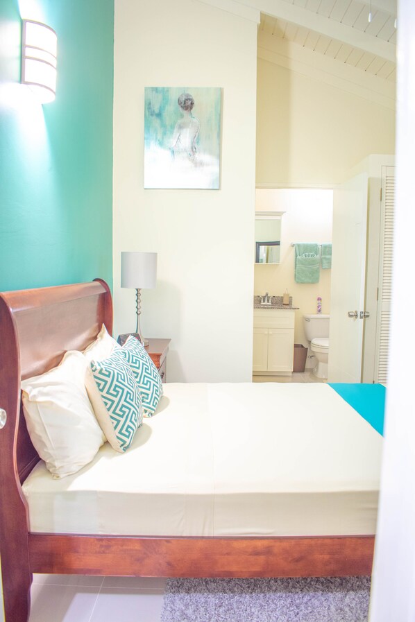 Une chambre avec un lit en bois, une table de chevet avec une lampe, un tableau au mur et une vue sur la salle de bain.