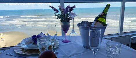 Una mesa servida con una botella de champán, copas, un jarrón con flores y vista a la playa.
