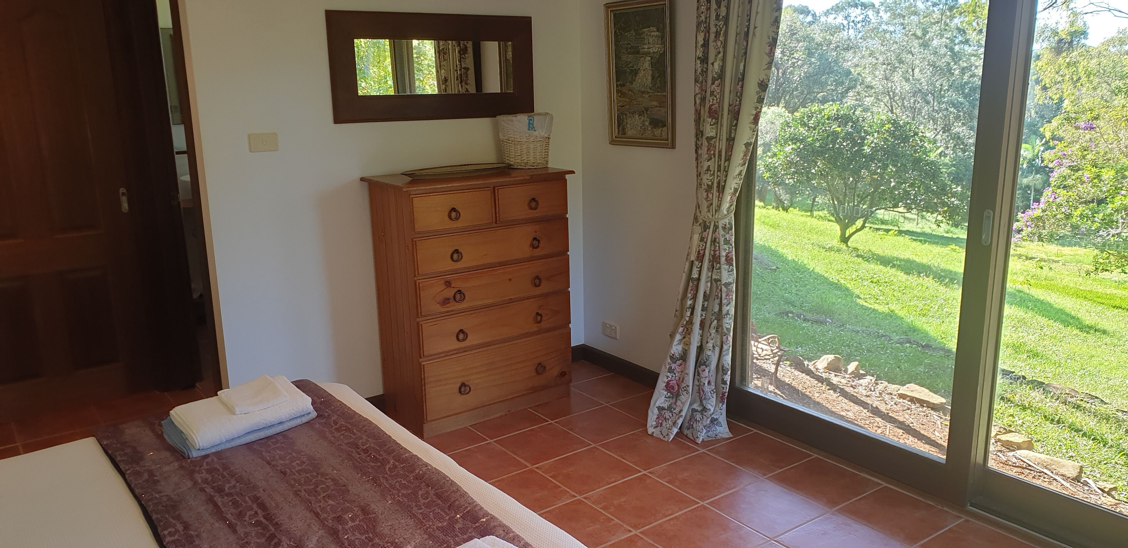 A bedroom with a wooden dresser, a bed with a red and white striped blanket, a window with a view of a garden, and a painting on the wall.