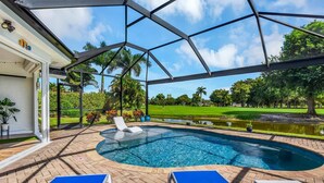 A covered swimming pool with lounge chairs, surrounded by a landscaped garden and a view of a golf course. - TORREY PINES - Single Family Pool Home in Lely Country Club (Naples)