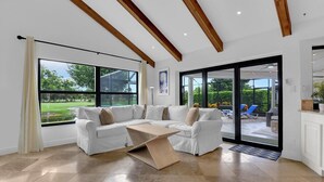 A spacious living room with a white sectional sofa, a wooden coffee table, and large windows offering a view of a garden and outdoor seating area. - TORREY PINES - Single Family Pool Home in Lely Country Club (Naples)