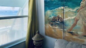 A room with a large window, a painting of a beach scene, and a small decorative item on the windowsill. - Oceanview Sealofts: NO spiral staircase & only 25 steps to ocean (Frigate Bay)