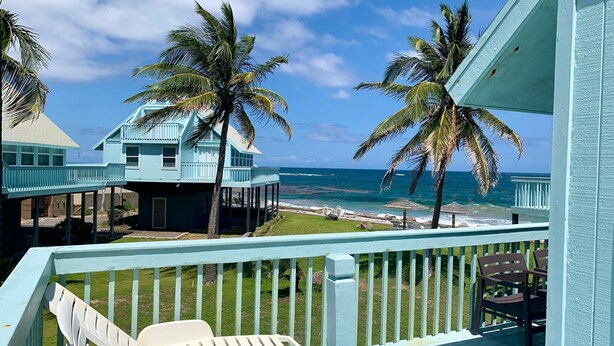 A beachfront property with a light blue house, a balcony with white chairs, and palm trees. - Oceanview Sealofts: NO spiral staircase & only 25 steps to ocean (Frigate Bay)