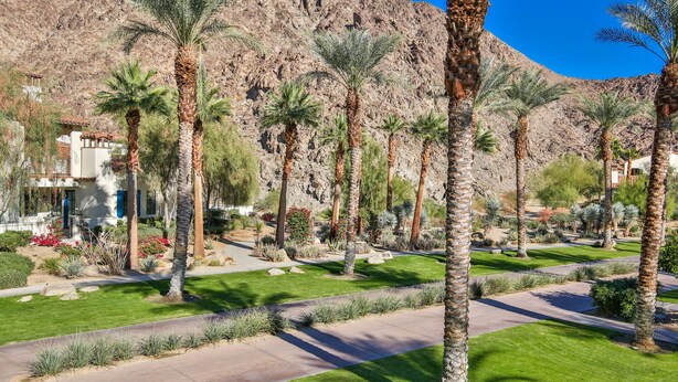 A landscaped garden with palm trees, a pathway, and a building with a tiled roof. - Mountain Views! Mountains, Palm Trees and Blue Skies - Upstairs with Garage, Pet Friendly (L56) (La Quinta)