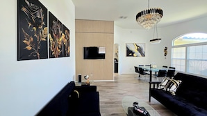 Ein modernes Wohnzimmer mit einer schwarzen Couch, einem Glastisch und einem Kronleuchter. An der Wand befinden sich drei schwarz-goldene Gemälde und ein an einer Holzvertäfelung befestigtes Fernsehgerät.