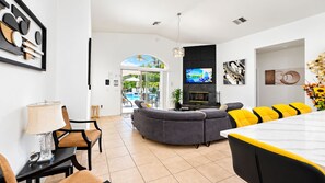 A modern living room with a curved grey sofa, yellow accent chairs, a dining table, and a TV mounted on a black panel. - The Panorama Oasis: Saltwater Pool, Spa & EV Charger (Cathedral City)