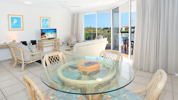 Un vaste espace de vie comprenant un coin repas avec une table à plateau en verre entourée de chaises en osier, un canapé et un téléviseur posé sur un meuble en bois.