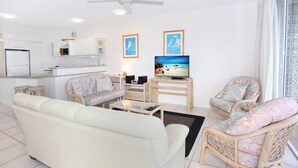Un salon comprenant un canapé, des fauteuils, une table basse, un téléviseur, ainsi qu’un coin cuisine avec des placards et un réfrigérateur.