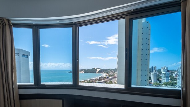A room with a curved bay window overlooking a coastal cityscape, including a tall building and a beach. - A retreat with a perfect sea view, 2 bedrooms, Wi-Fi, 24-hour concierge, rotating garage. (Natal)