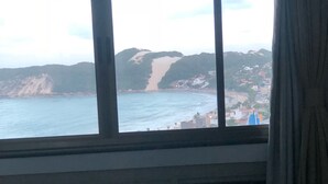 A room with a bed, a window with a view of a beach and hills, and a curtain on the window. - A retreat with a perfect sea view, 2 bedrooms, Wi-Fi, 24-hour concierge, rotating garage. (Natal)