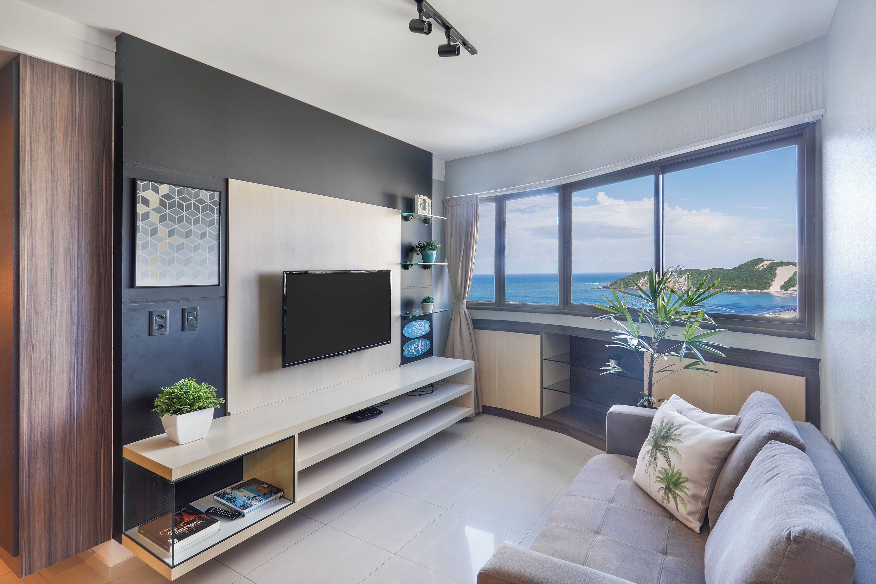 A modern living room with a flat-screen TV, a sofa, a coffee table, and a large window offering a view of the sea.