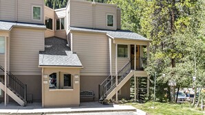 A multi-story building with a staircase leading to a balcony, surrounded by trees and a hilly background. - Entire 1BR apt, Sleeps 4, Wi-Fi, Close to Slopes (Vail)
