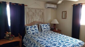 Een slaapkamer met een bed, gordijnen, een plafondventilator, een airconditioning, een bedlampje, een klein tafeltje met een vaas, en een macramé wanddecoratie.