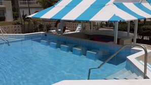 Een zwembad met een blauw-wit gestreepte parasol, een liggedeelte aan de rand en een reddingsboei.