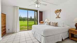 Un dormitorio con una cama grande, un ventilador de techo, vista a un paisaje verde a través de una puerta corrediza y una mesita de noche de mimbre.