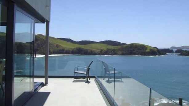 A balcony with a glass railing overlooking a coastal landscape with hills and a body of water.