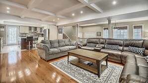 Uma sala de estar ampla com um grande sofá de couro marrom, um sofá cinza e uma mesa de centro de madeira. Dá para ver uma cozinha ao fundo, com uma área de jantar e uma escada que leva para o andar superior.