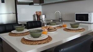 A modern kitchen with a white countertop, a stainless steel refrigerator, a microwave, and a sink. There are woven placemats under bowls and plates, and a glass of orange juice on the counter. - Phaedrus Living: Seaside Luxury Flat Lighthouse 53 (Paphos)