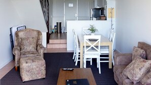 Een woonkamer met een bank, fauteuil en een kleine tafel met een boek. Een trap leidt naar een keuken met een eettafel en stoelen.