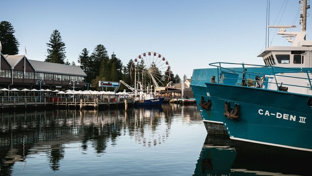 Een haven met een grote blauwe boot genaamd CA-DEN III, een reuzenrad en een restaurant aan het water.