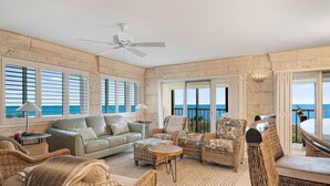 A spacious living area with a view of the ocean, furnished with wicker chairs, a sofa, and a ceiling fan. - Land`s End Beachfront Elegance: Top Floor Corner (Treasure Island)