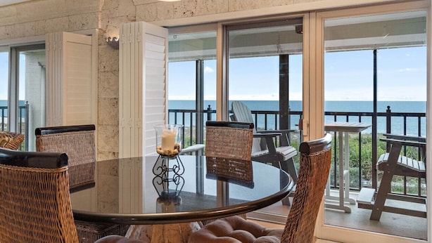 A round dining table with wicker chairs, a candle centerpiece, and a view of the ocean through large windows. - Land`s End Beachfront Elegance: Top Floor Corner (Treasure Island)