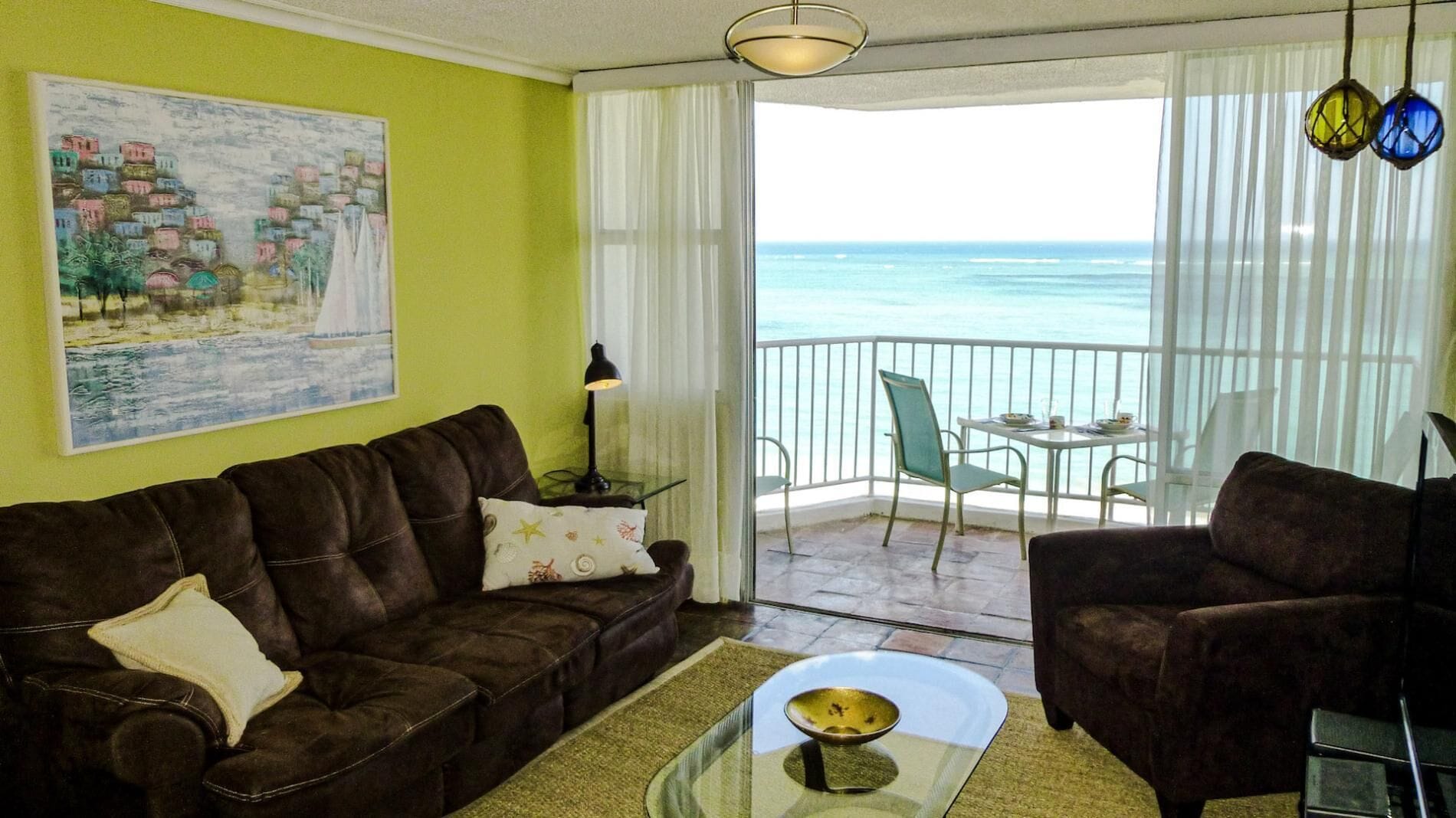 Un salon avec un canapé en cuir marron, une table basse en verre et un tableau représentant un paysage côtier. Il y a un balcon avec une table et des chaises, d’où l’on a vue sur la mer.